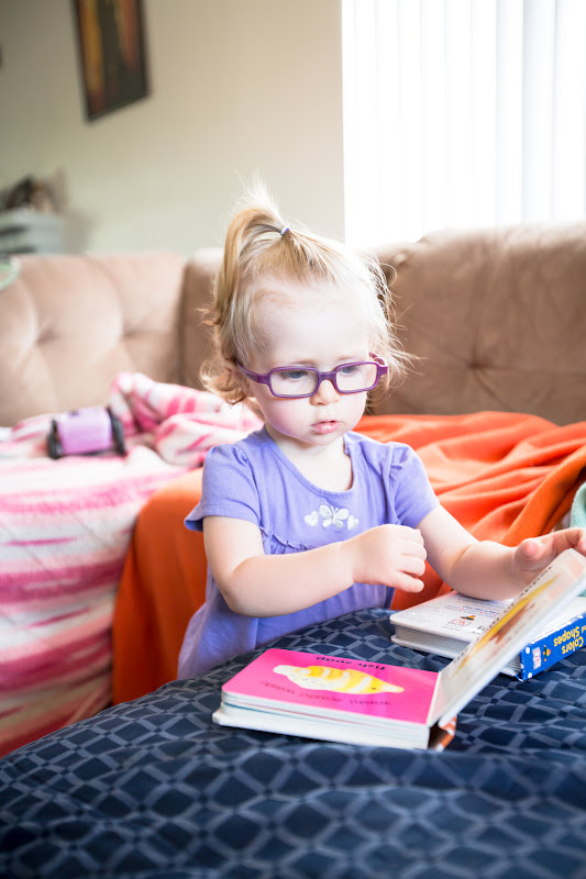 16 month old girl with a purple shirt and dark red glasses reading a book
