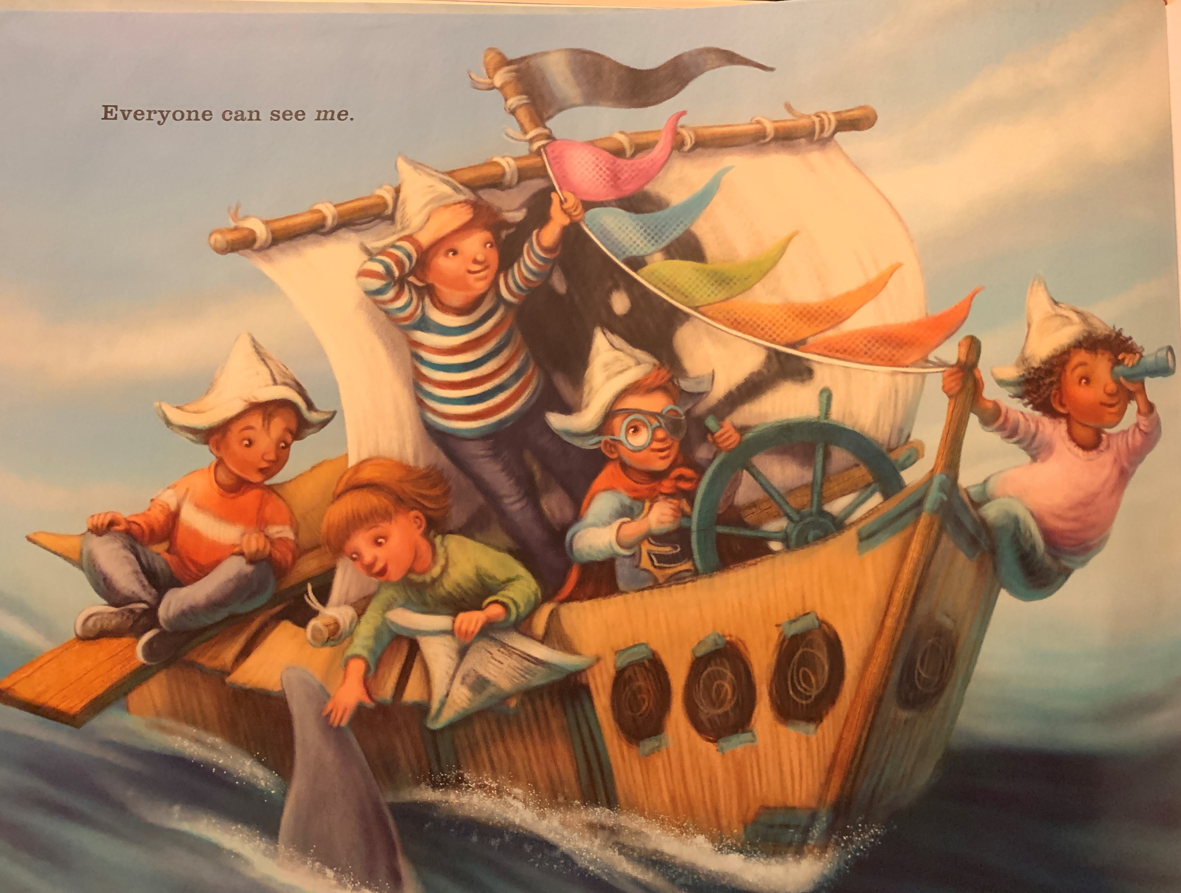 Page from the book.  Five children are riding on a ship with colorful flags. One girl is petting a dolphin.  The text on the page reads, "Everyone can see me."
