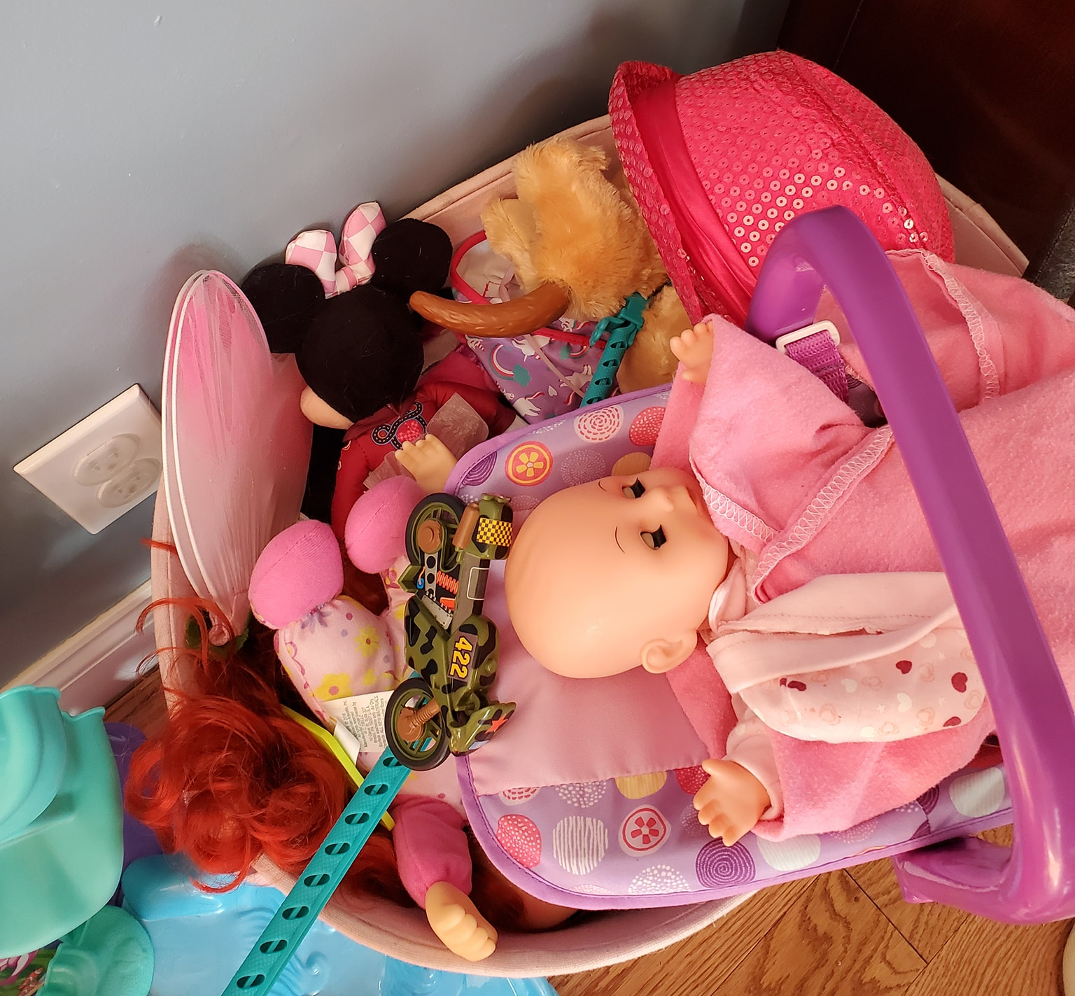 photo of a bin of a child's toys, including dolls, a hat, a motorcyle, dress up fairy wings, and a pair of children's glasses