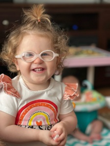 toddler girl with blond curls wearing pale pink glasses. She has a big smile